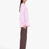 A woman modeling a light pink long-sleeve t-shirt with vertical blue text on the sleeve, paired with brown wide-leg pants and sunglasses, shown from a side profile against a light gray background.