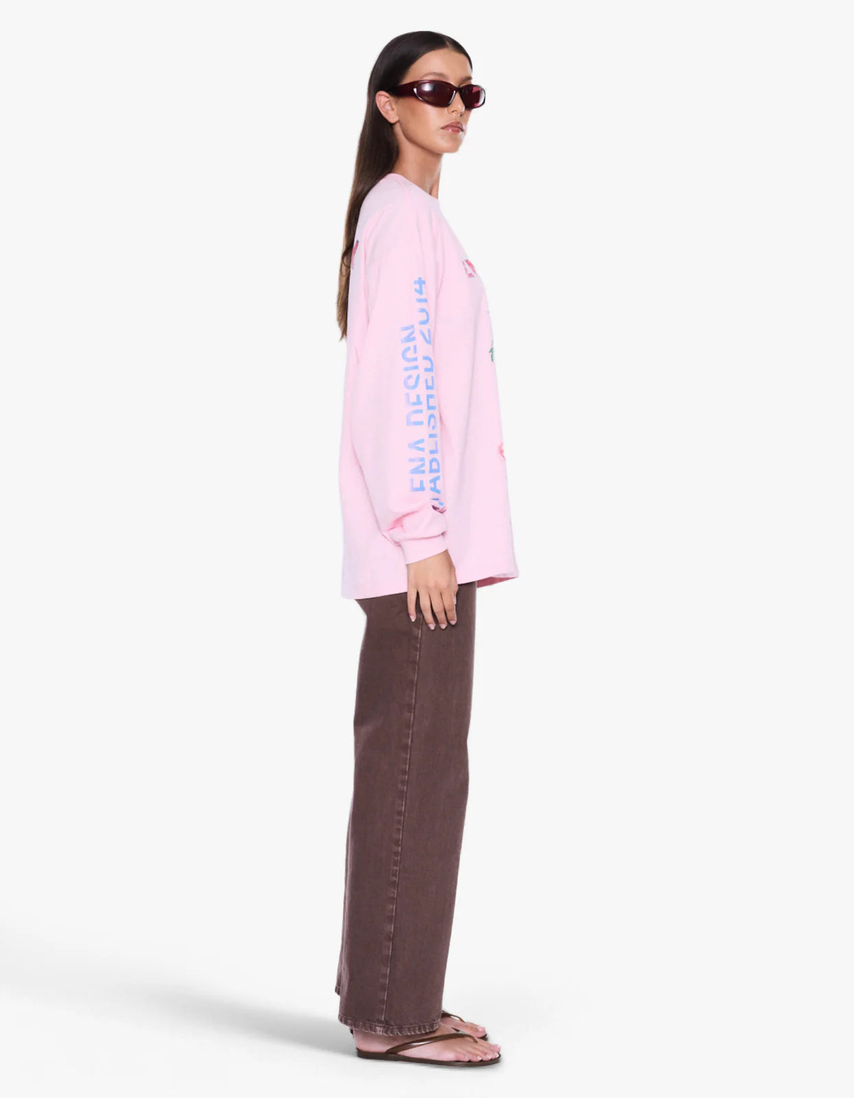 A woman modeling a light pink long-sleeve t-shirt with vertical blue text on the sleeve, paired with brown wide-leg pants and sunglasses, shown from a side profile against a light gray background.