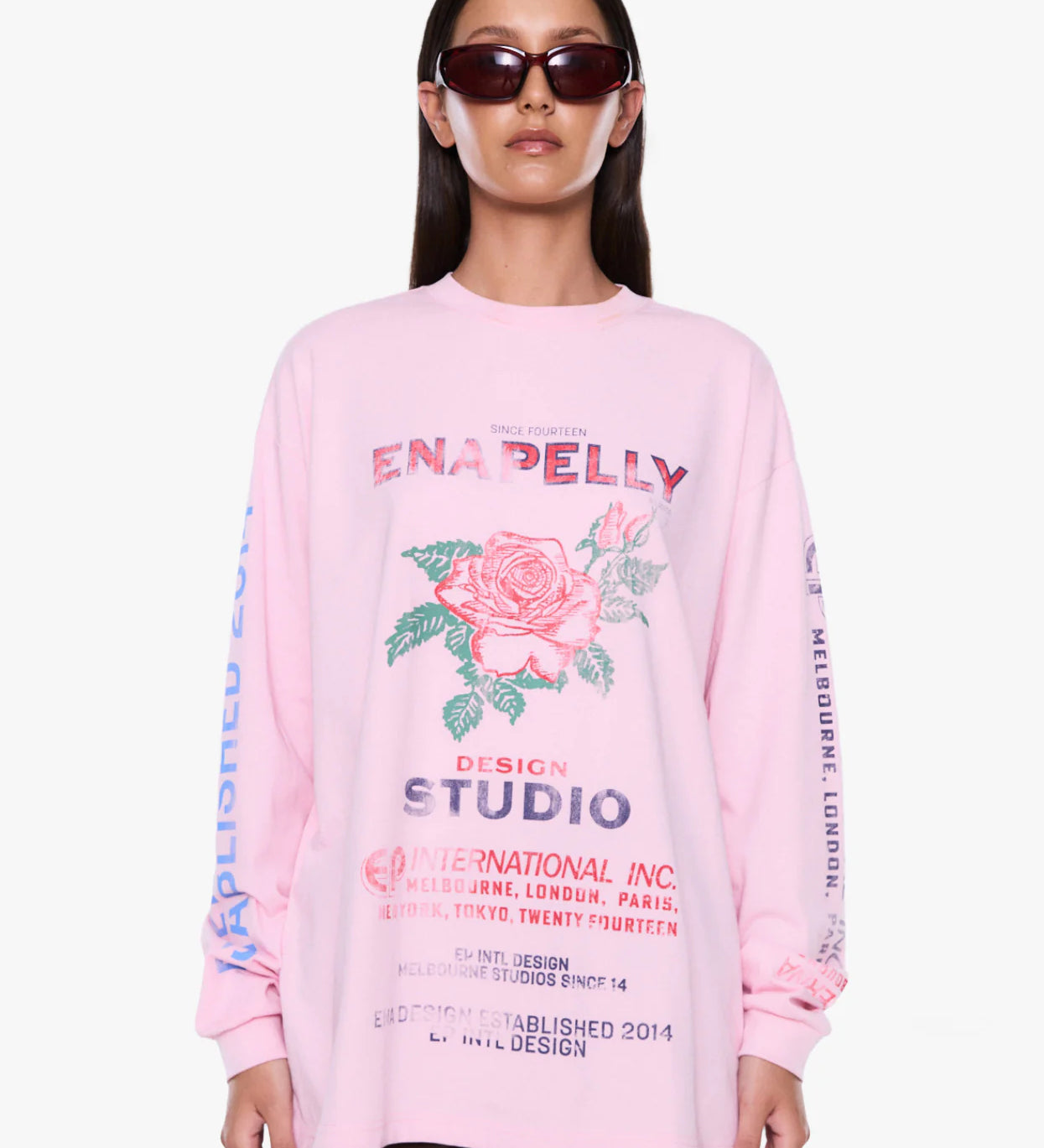 A model wearing a light pink long-sleeve graphic sweatshirt with a rose print and text, styled with dark sunglasses against a plain white background.
