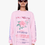 A model wearing a light pink long-sleeve graphic sweatshirt with a rose print and text, styled with dark sunglasses against a plain white background.