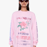 A model wearing a light pink long-sleeve graphic sweatshirt with a rose print and text, styled with dark sunglasses against a plain white background.