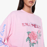 A close-up of a model wearing a light pink Enapellu crewneck sweatshirt with a large rose graphic and text on the front, set against a plain white background.