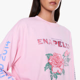 A close-up of a model wearing a light pink Enapellu crewneck sweatshirt with a large rose graphic and text on the front, set against a plain white background.
