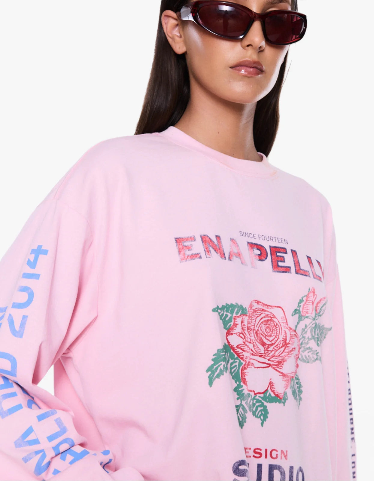 A close-up of a model wearing a light pink Enapellu crewneck sweatshirt with a large rose graphic and text on the front, set against a plain white background.
