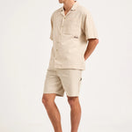 A man modeling a light beige short-sleeve camp shirt and matching shorts, paired with white socks and gray athletic sneakers, against a plain white studio background.