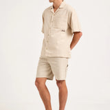A man modeling a light beige short-sleeve camp shirt and matching shorts, paired with white socks and gray athletic sneakers, against a plain white studio background.