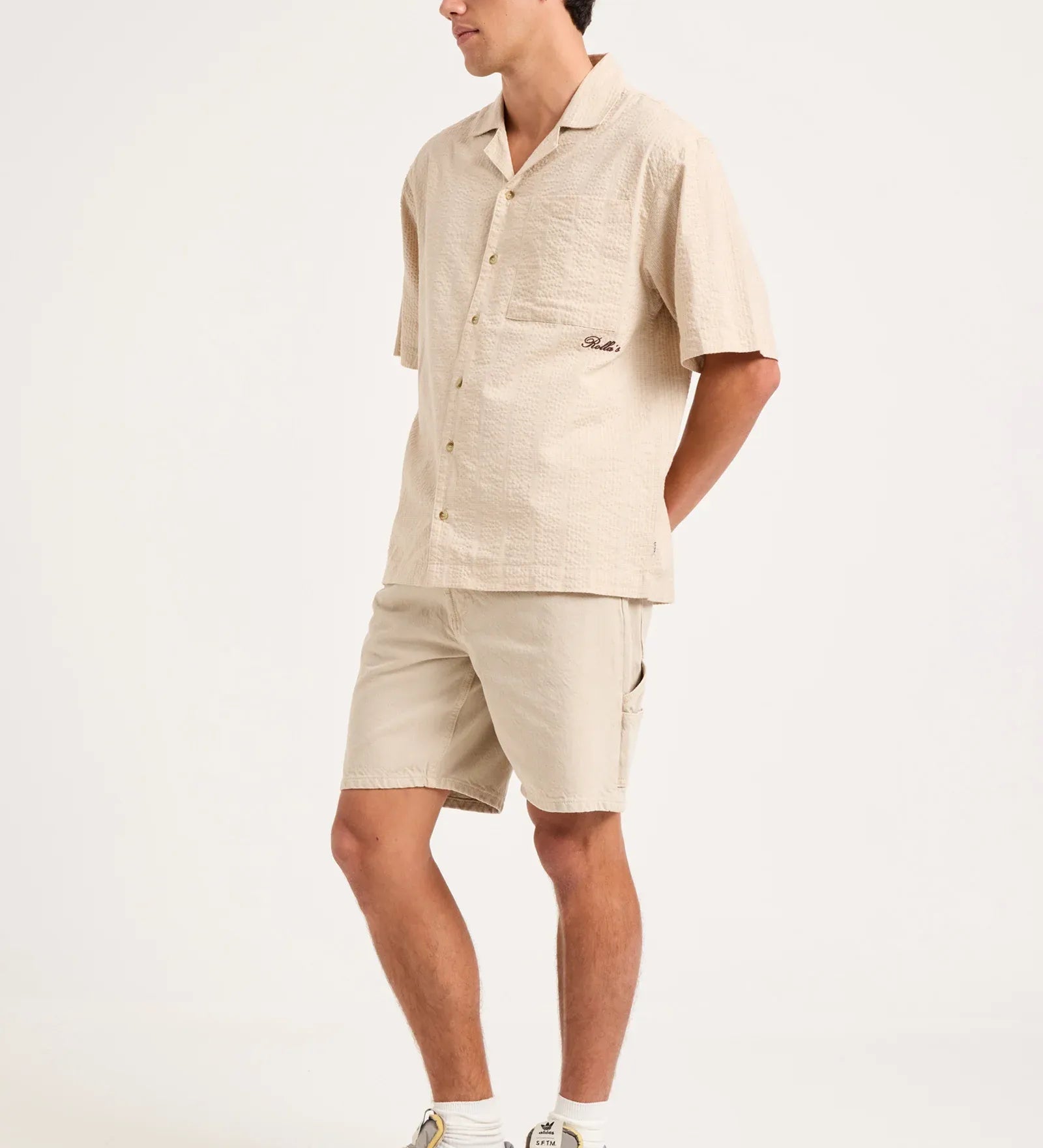 A man modeling a light beige short-sleeve camp shirt and matching shorts, paired with white socks and gray athletic sneakers, against a plain white studio background.