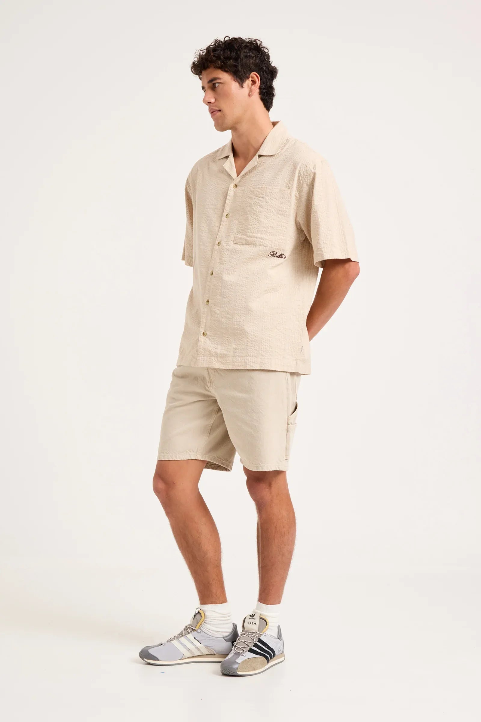 A man modeling a light beige short-sleeve camp shirt and matching shorts, paired with white socks and gray athletic sneakers, against a plain white studio background.