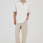 A man modeling a white short-sleeve button-up shirt with black and white patterned collar and sleeve cuffs, paired with beige trousers and gray sneakers, against a plain light gray studio background.
