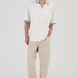 A man modeling a white short-sleeve button-up shirt with black and white patterned collar and sleeve cuffs, paired with beige trousers and gray sneakers, against a plain light gray studio background.