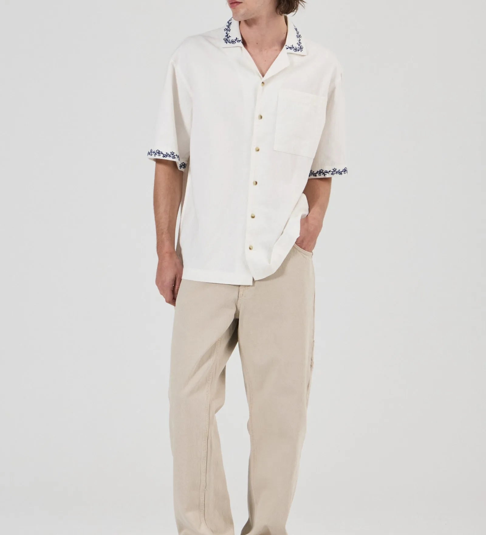 A man modeling a white short-sleeve button-up shirt with black and white patterned collar and sleeve cuffs, paired with beige trousers and gray sneakers, against a plain light gray studio background.