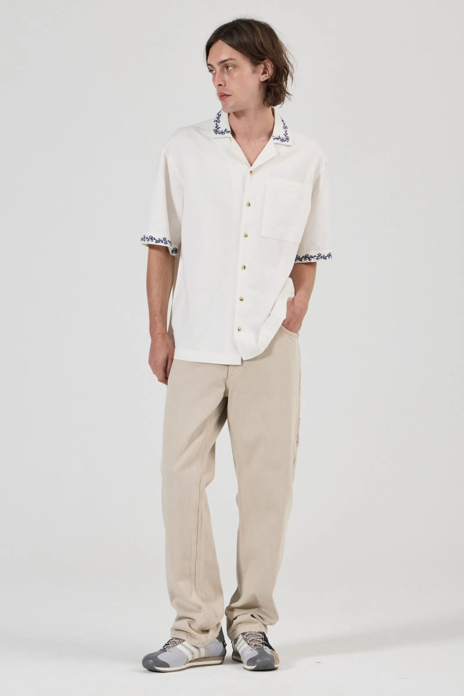 A man modeling a white short-sleeve button-up shirt with black and white patterned collar and sleeve cuffs, paired with beige trousers and gray sneakers, against a plain light gray studio background.