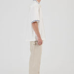 A model shown in profile wearing a white short-sleeve polo shirt with black and white striped trim on the collar and sleeve cuffs, paired with beige trousers and gray sneakers, against a plain light gray studio background.