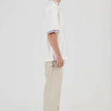 A model shown in profile wearing a white short-sleeve polo shirt with black and white striped trim on the collar and sleeve cuffs, paired with beige trousers and gray sneakers, against a plain light gray studio background.