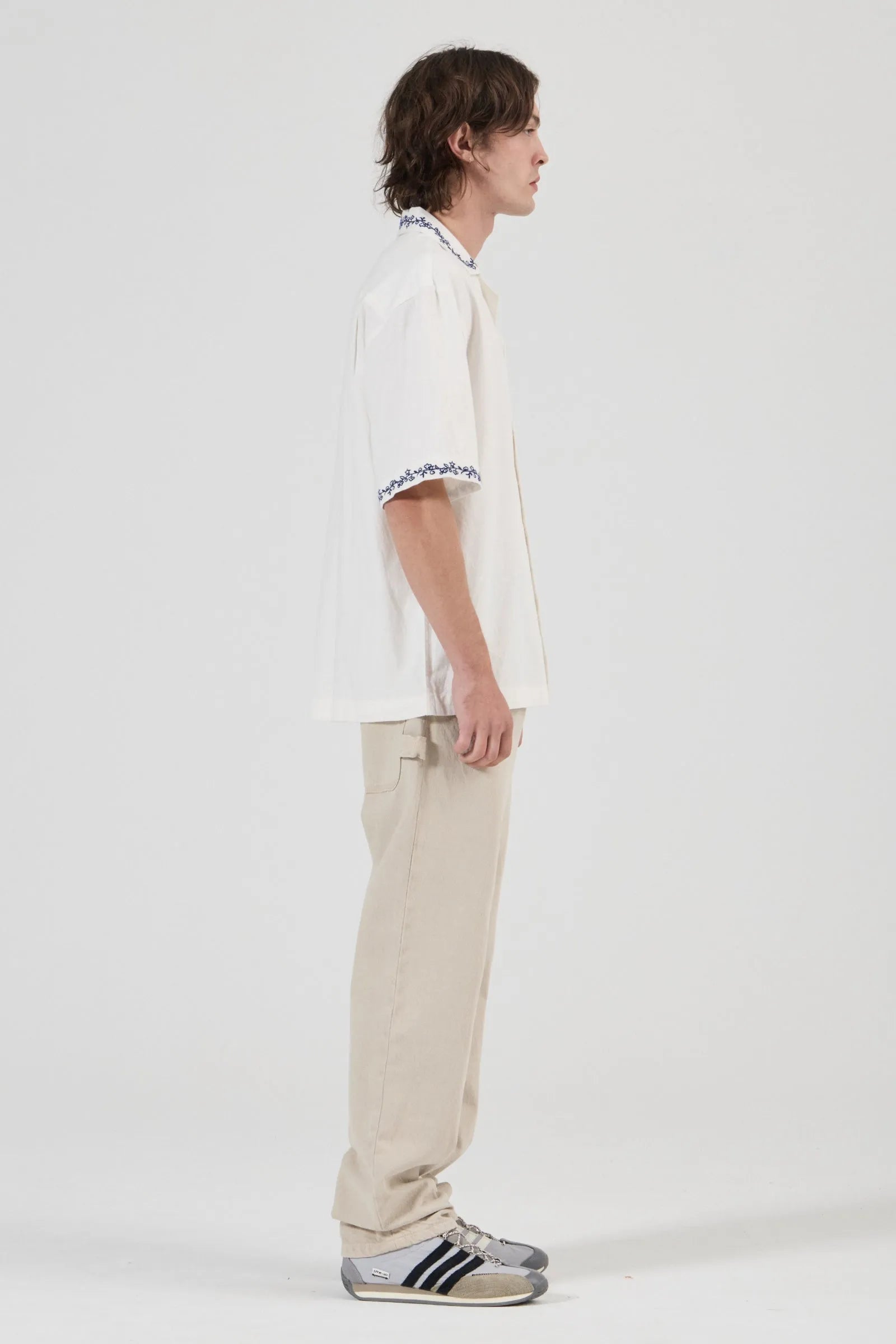 A model shown in profile wearing a white short-sleeve polo shirt with black and white striped trim on the collar and sleeve cuffs, paired with beige trousers and gray sneakers, against a plain light gray studio background.