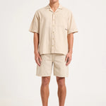 A man modeling a light beige short-sleeve button-up shirt and matching shorts set, paired with gray sneakers, against a plain light gray studio background.