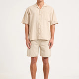 A man modeling a light beige short-sleeve button-up shirt and matching shorts set, paired with gray sneakers, against a plain light gray studio background.
