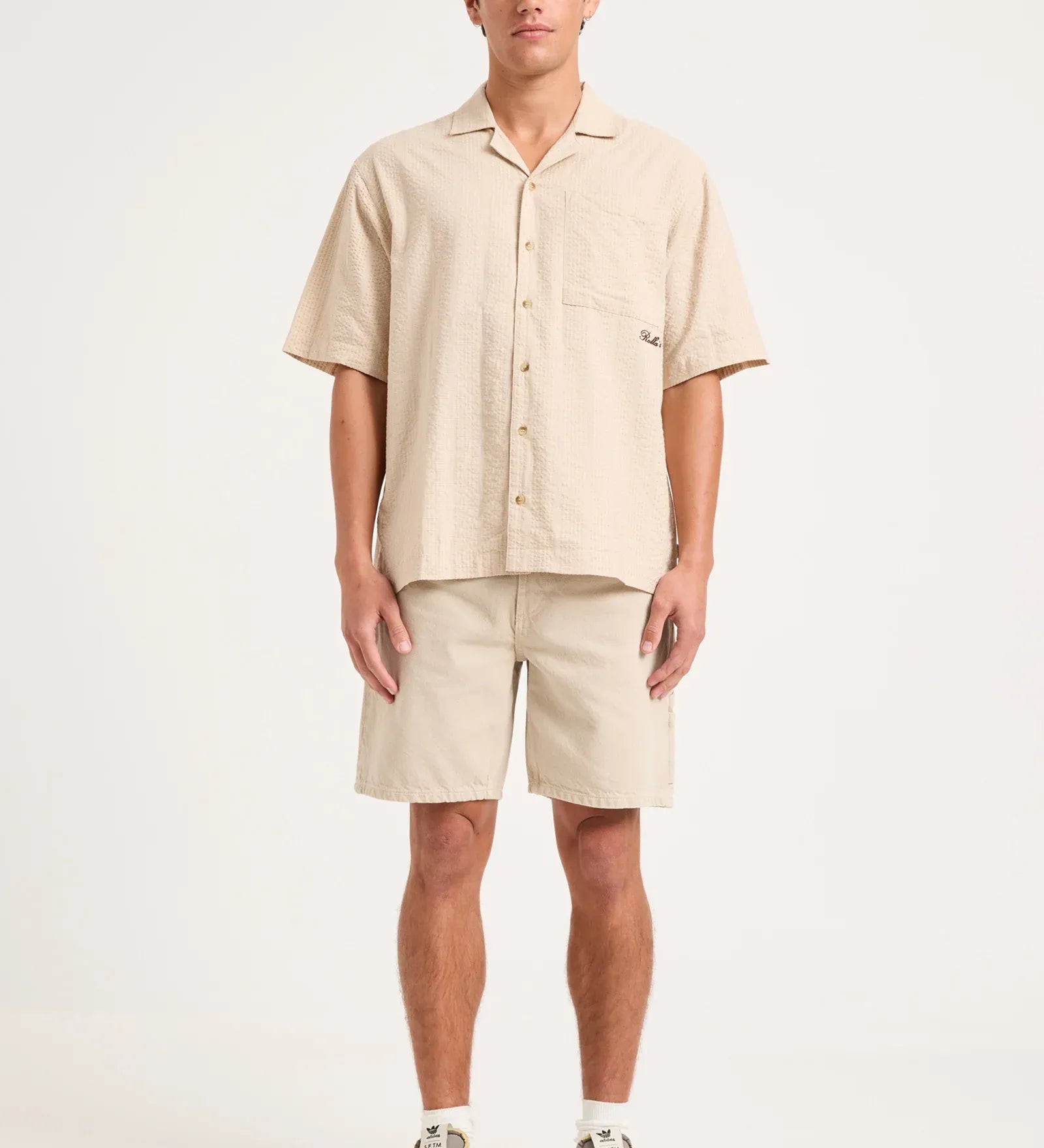 A man modeling a light beige short-sleeve button-up shirt and matching shorts set, paired with gray sneakers, against a plain light gray studio background.