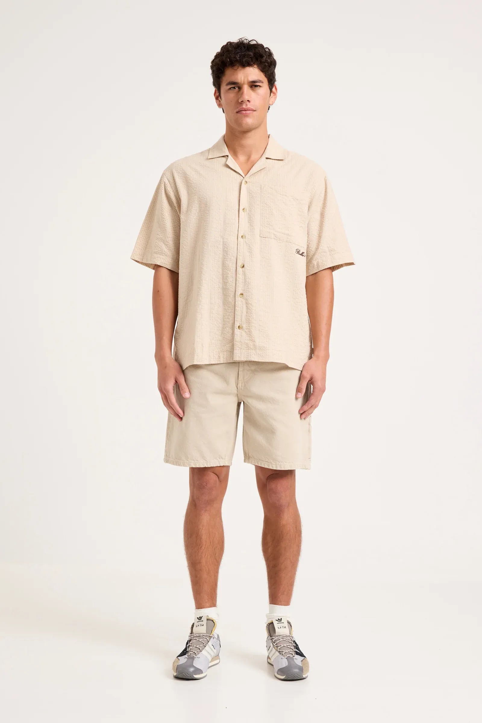 A man modeling a light beige short-sleeve button-up shirt and matching shorts set, paired with gray sneakers, against a plain light gray studio background.