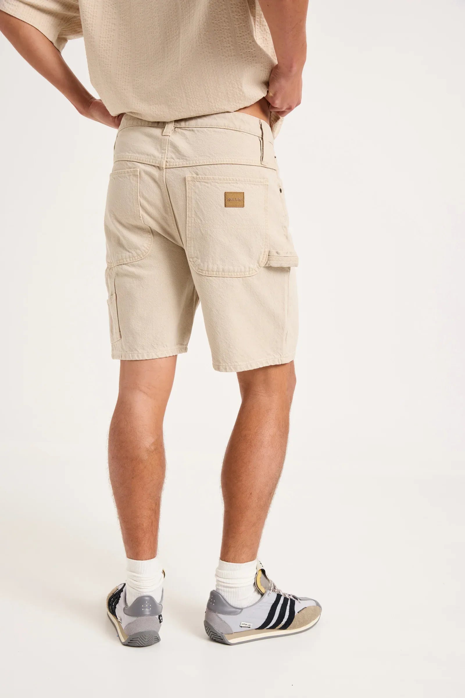 A person shown from the back wearing light beige cargo shorts, a matching beige top, white socks, and gray sneakers against a plain white studio background.