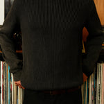 Person wearing a dark ribbed sweater standing in front of a bookshelf with books.