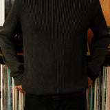 Person wearing a dark ribbed sweater standing in front of a bookshelf with books.