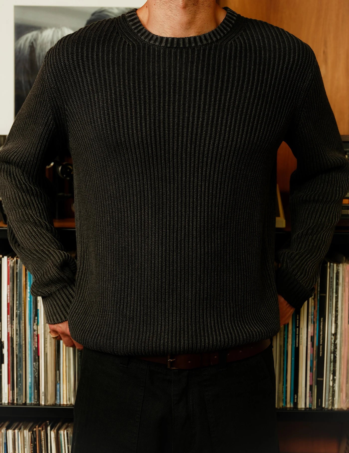 Person wearing a dark ribbed sweater standing in front of a bookshelf with books.