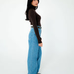 A woman stands in profile against a plain white studio background, wearing a fitted dark brown long-sleeve turtleneck top, light blue wide-leg jeans, and black shoes.