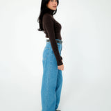 A woman stands in profile against a plain white studio background, wearing a fitted dark brown long-sleeve turtleneck top, light blue wide-leg jeans, and black shoes.