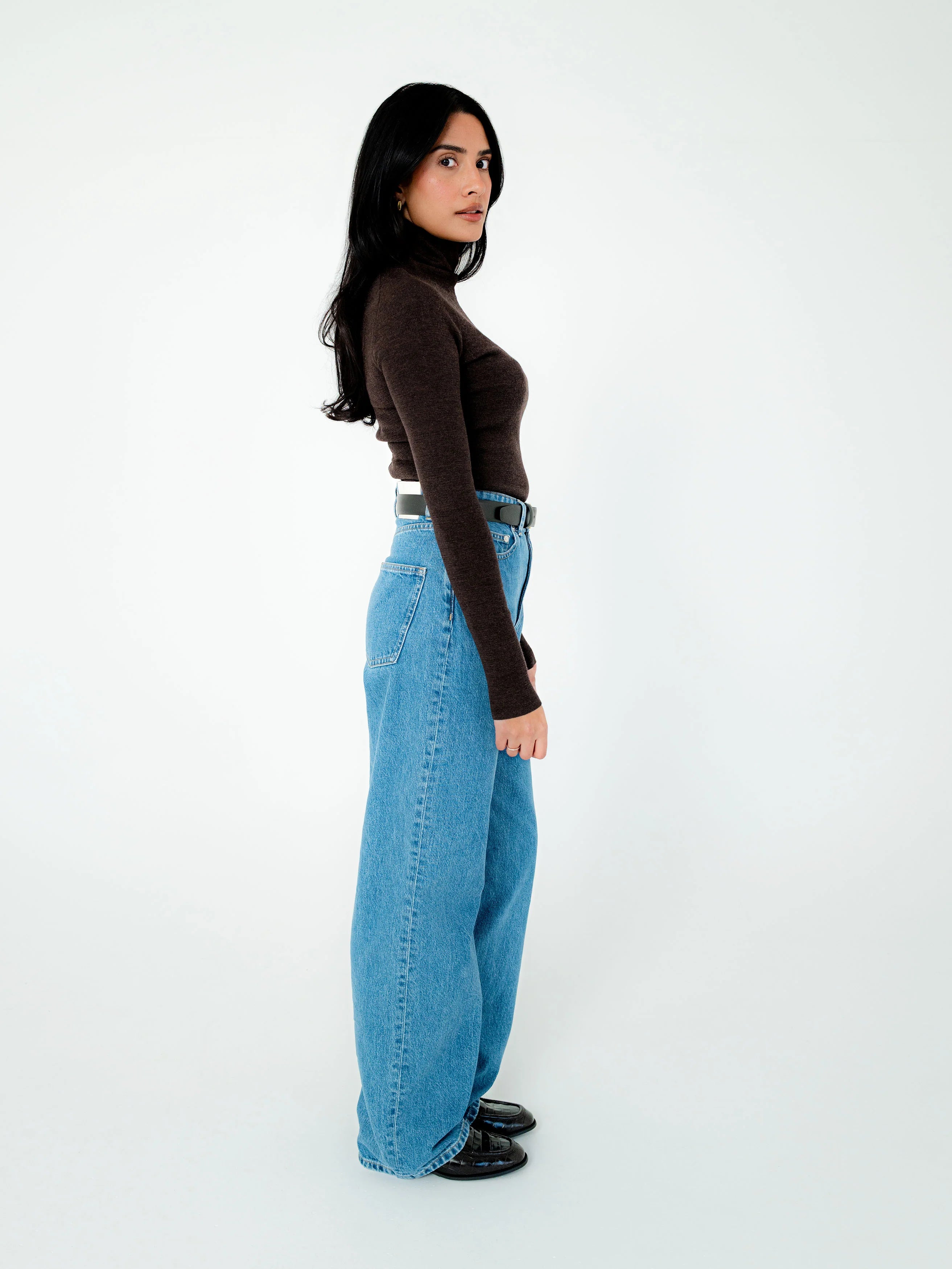 A woman stands in profile against a plain white studio background, wearing a fitted dark brown long-sleeve turtleneck top, light blue wide-leg jeans, and black shoes.