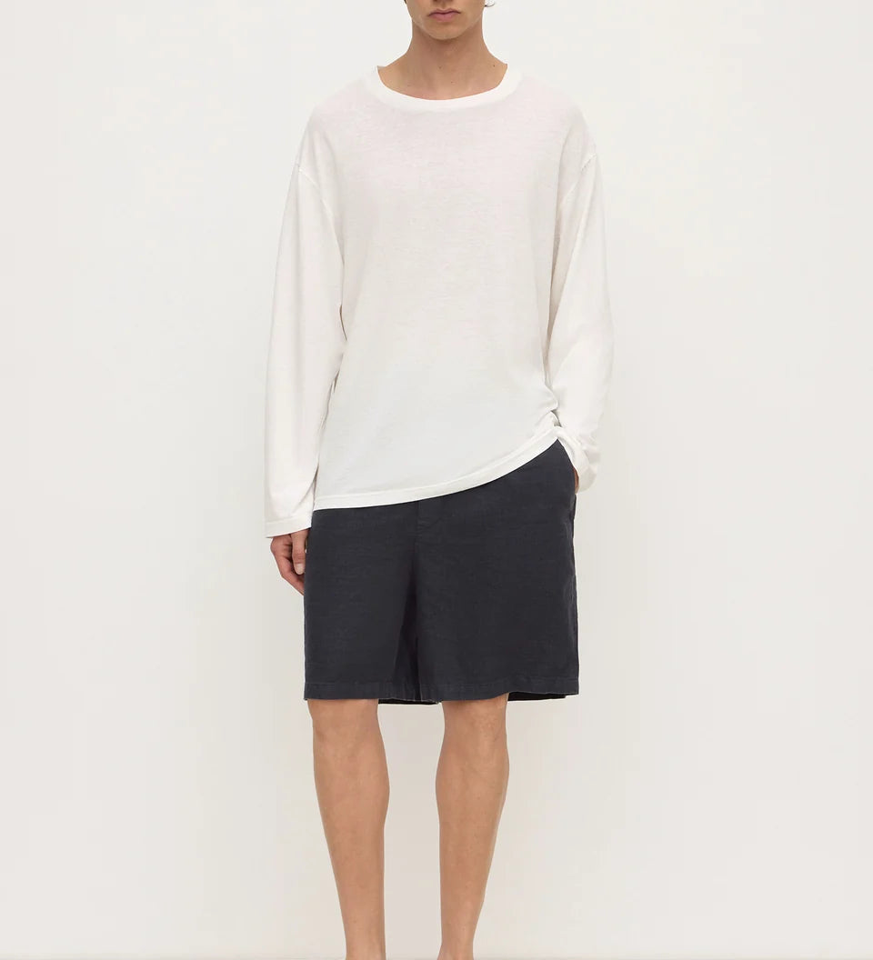 A male model stands against a plain white studio background wearing a loose white long-sleeve knit top, dark charcoal shorts, and black slip-on loafers.