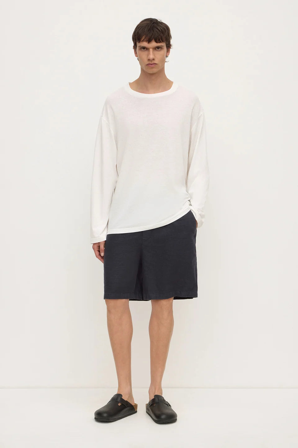 A male model stands against a plain white studio background wearing a loose white long-sleeve knit top, dark charcoal shorts, and black slip-on loafers.