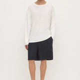 A male model stands against a plain white studio background wearing a loose white long-sleeve knit top, dark charcoal shorts, and black slip-on loafers.