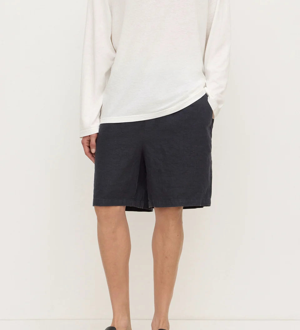 A model wearing a white long-sleeve knit top, dark charcoal gray shorts, and black slip-on clogs, standing against a plain off-white studio background.
