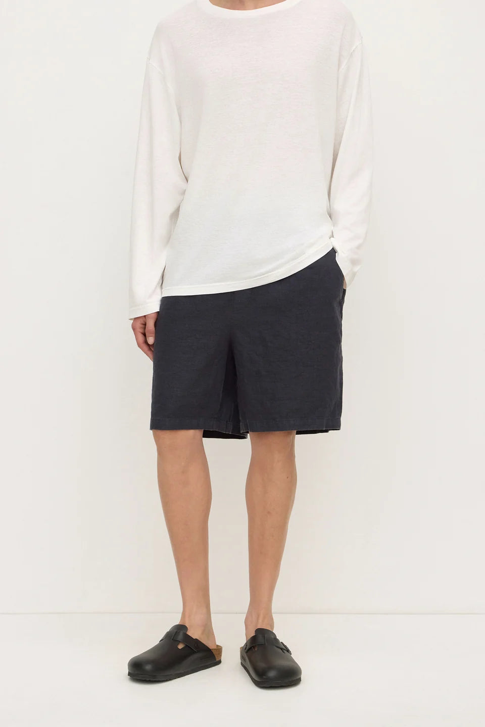 A model wearing a white long-sleeve knit top, dark charcoal gray shorts, and black slip-on clogs, standing against a plain off-white studio background.