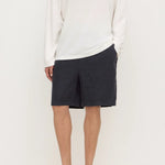 A model wearing a white long-sleeve knit top, dark charcoal gray shorts, and black slip-on clogs, standing against a plain off-white studio background.