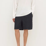 A model wearing a white long-sleeve knit top, dark charcoal gray shorts, and black slip-on clogs, standing against a plain off-white studio background.