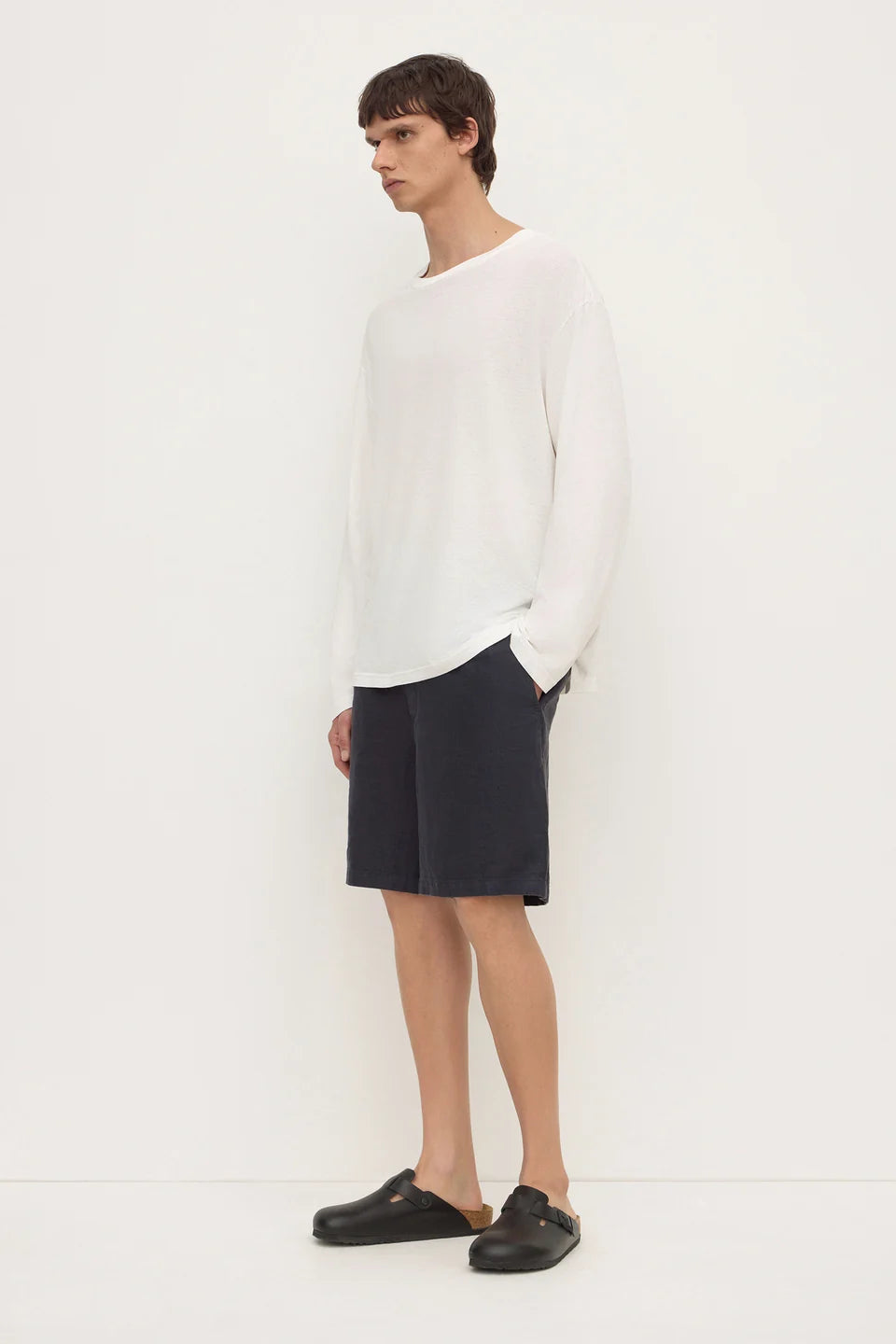 A man modeling a white long-sleeve knit sweater, dark navy shorts, and black clogs against a plain white studio background.