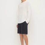 A man modeling a white long-sleeve knit sweater, dark navy shorts, and black clogs against a plain white studio background.