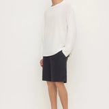 A man modeling a white long-sleeve knit sweater, dark navy shorts, and black clogs against a plain white studio background.