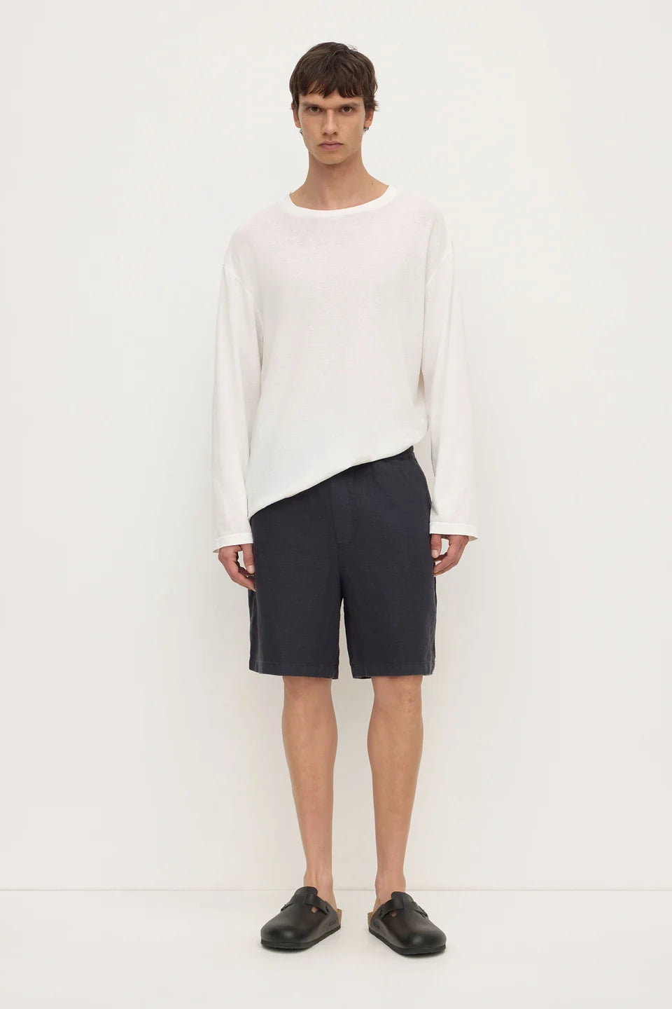 A male model stands against a plain white studio background wearing a loose white long-sleeve top, dark navy shorts, and black slip-on loafers.