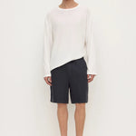 A male model stands against a plain white studio background wearing a loose white long-sleeve top, dark navy shorts, and black slip-on loafers.