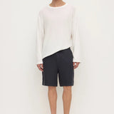 A male model stands against a plain white studio background wearing a loose white long-sleeve top, dark navy shorts, and black slip-on loafers.
