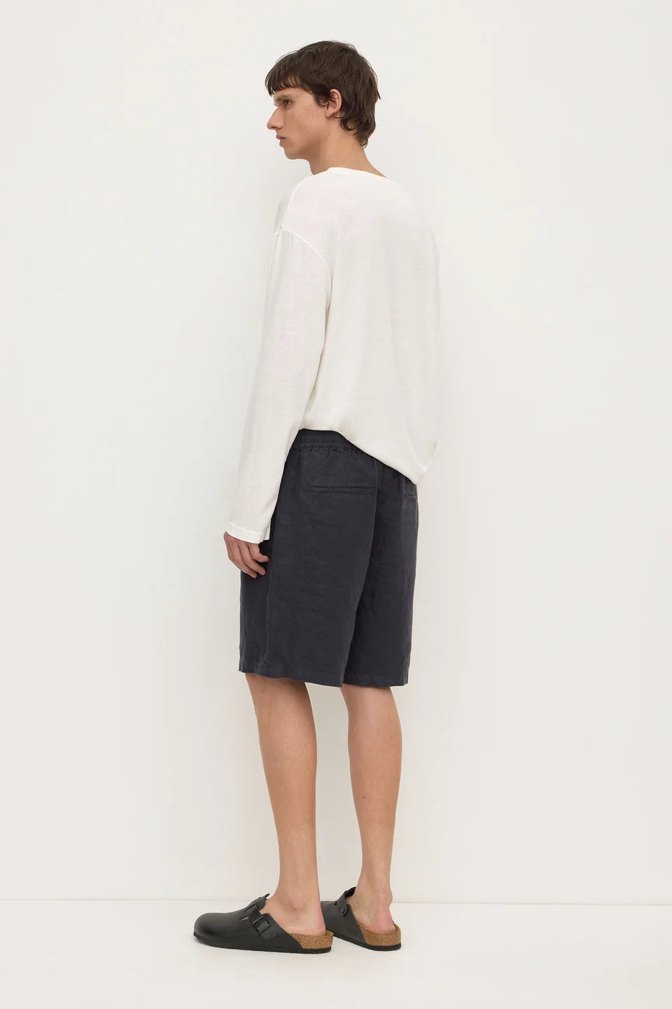 A man stands in profile wearing a loose white long-sleeve top, dark charcoal shorts, and dark slip-on shoes against a plain white studio background.