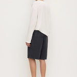 A man stands in profile wearing a loose white long-sleeve top, dark charcoal shorts, and dark slip-on shoes against a plain white studio background.