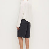 A man stands in profile wearing a loose white long-sleeve top, dark charcoal shorts, and dark slip-on shoes against a plain white studio background.