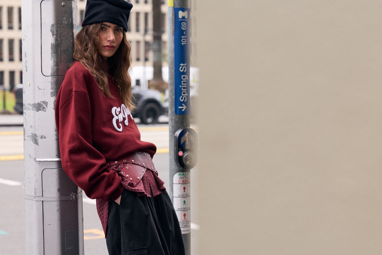 A woman wearing a burgundy sweatshirt with a white graphic, a black beret, and a black skirt leans against a city street pole.