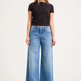 A woman modeling a pair of wide-leg light blue denim jeans with a black short-sleeve t-shirt, standing against a plain white studio background.
