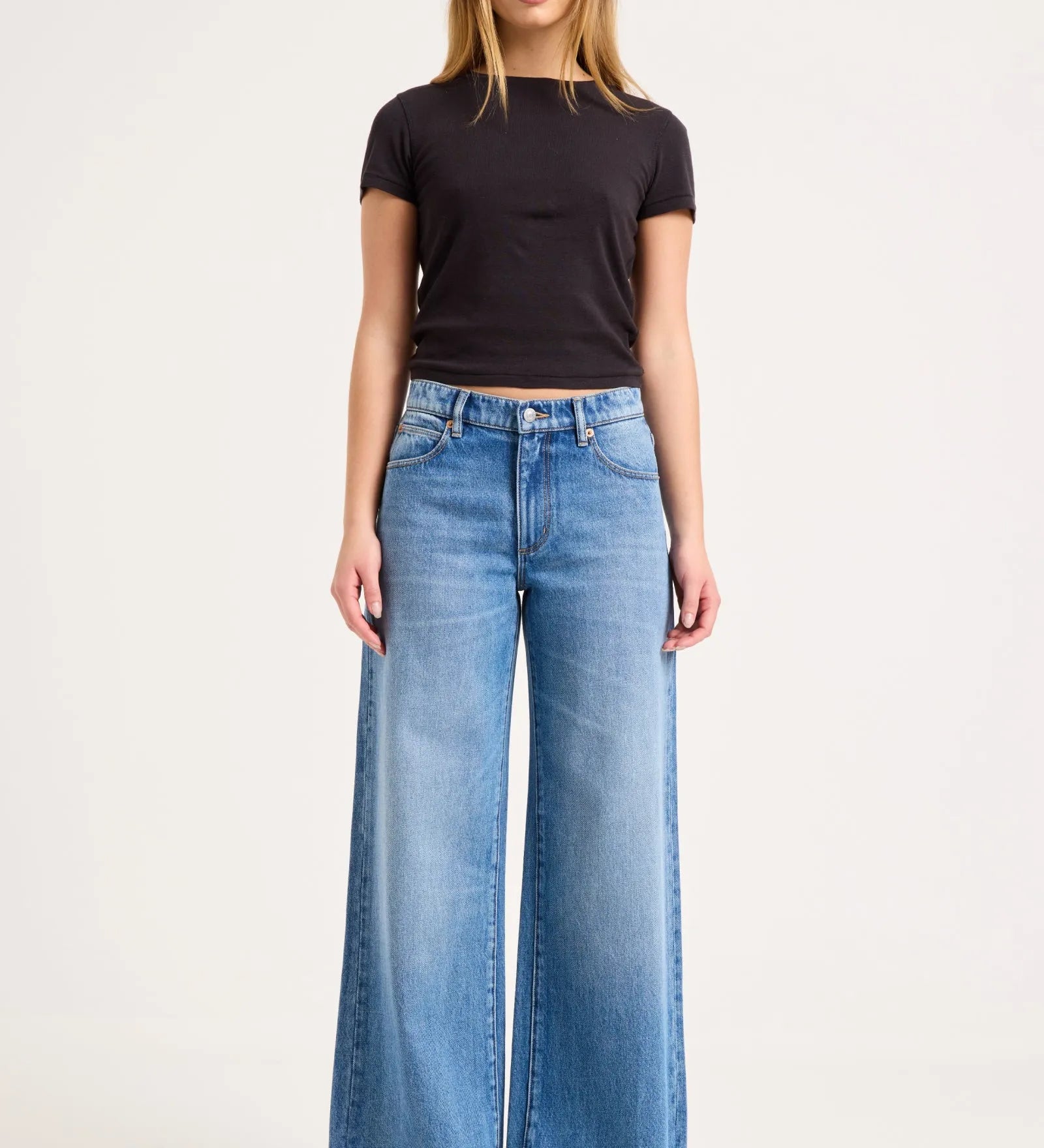 A woman modeling a pair of wide-leg light blue denim jeans with a black short-sleeve t-shirt, standing against a plain white studio background.