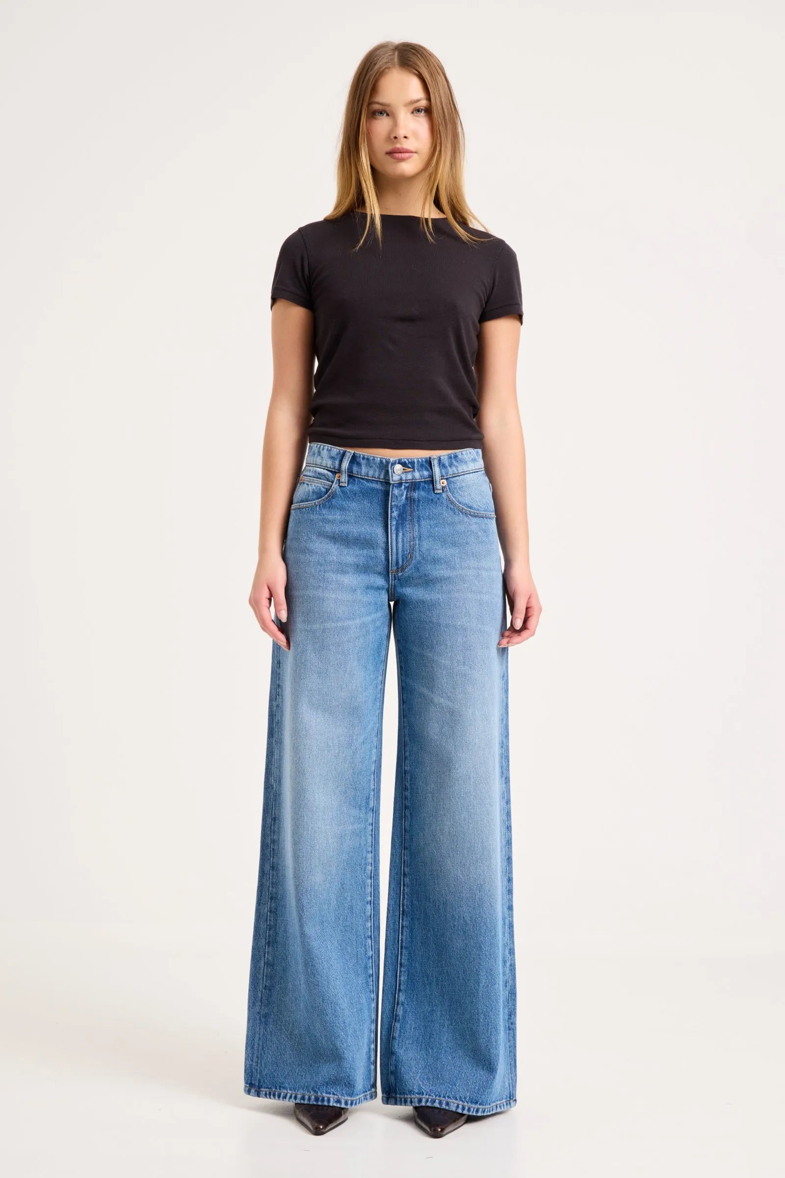 A woman modeling a pair of wide-leg light blue denim jeans with a black short-sleeve t-shirt, standing against a plain white studio background.